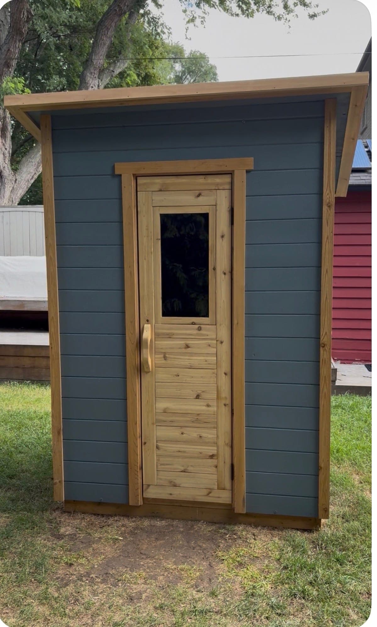The 2025 portable sauna outdoors: dark blue-grey horizontal siding, natural cedar corner boards and a narrow handmade door with a tall window, under a single-slope roof