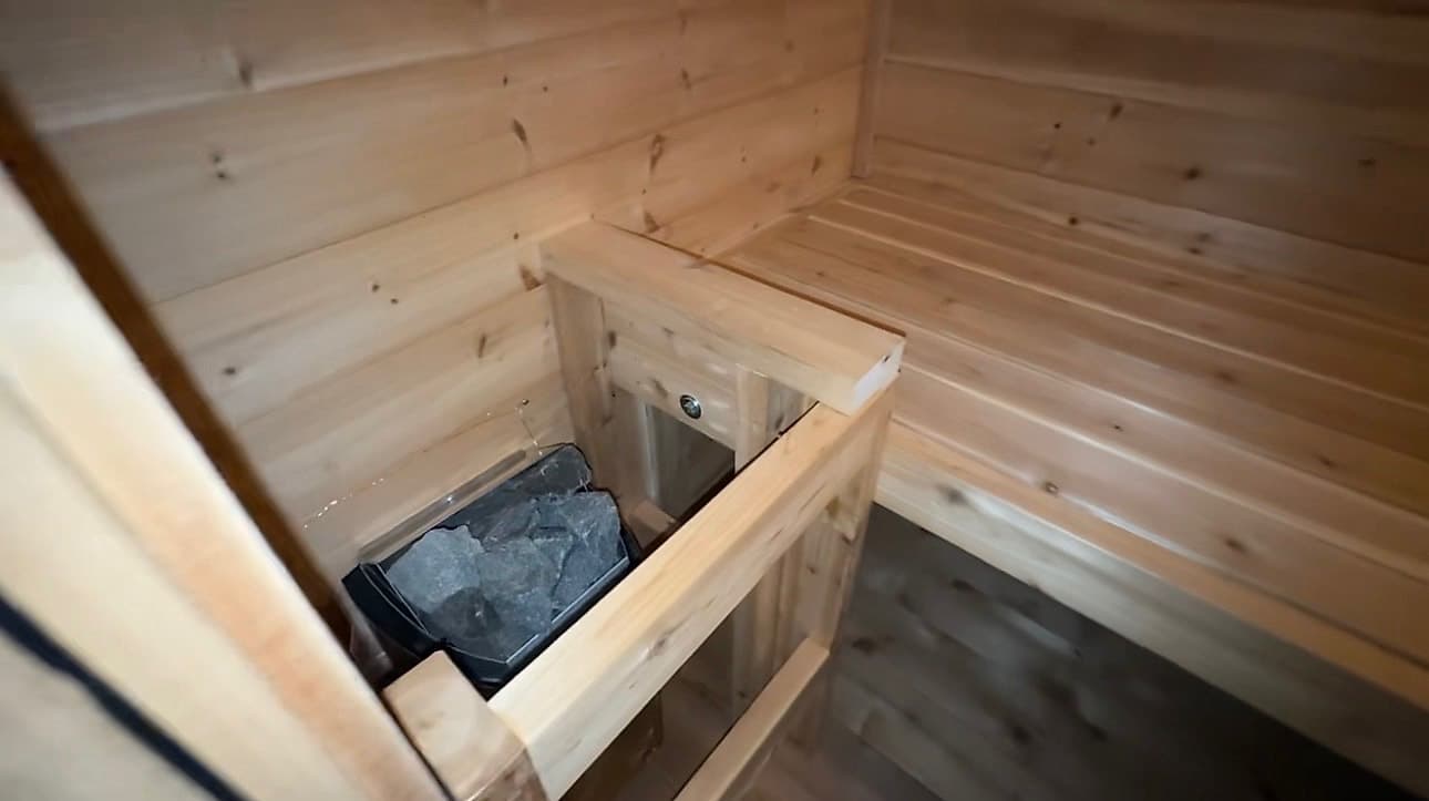 The heater and its stones inside the portable sauna, enclosed by a wooden guard rail beside the bench