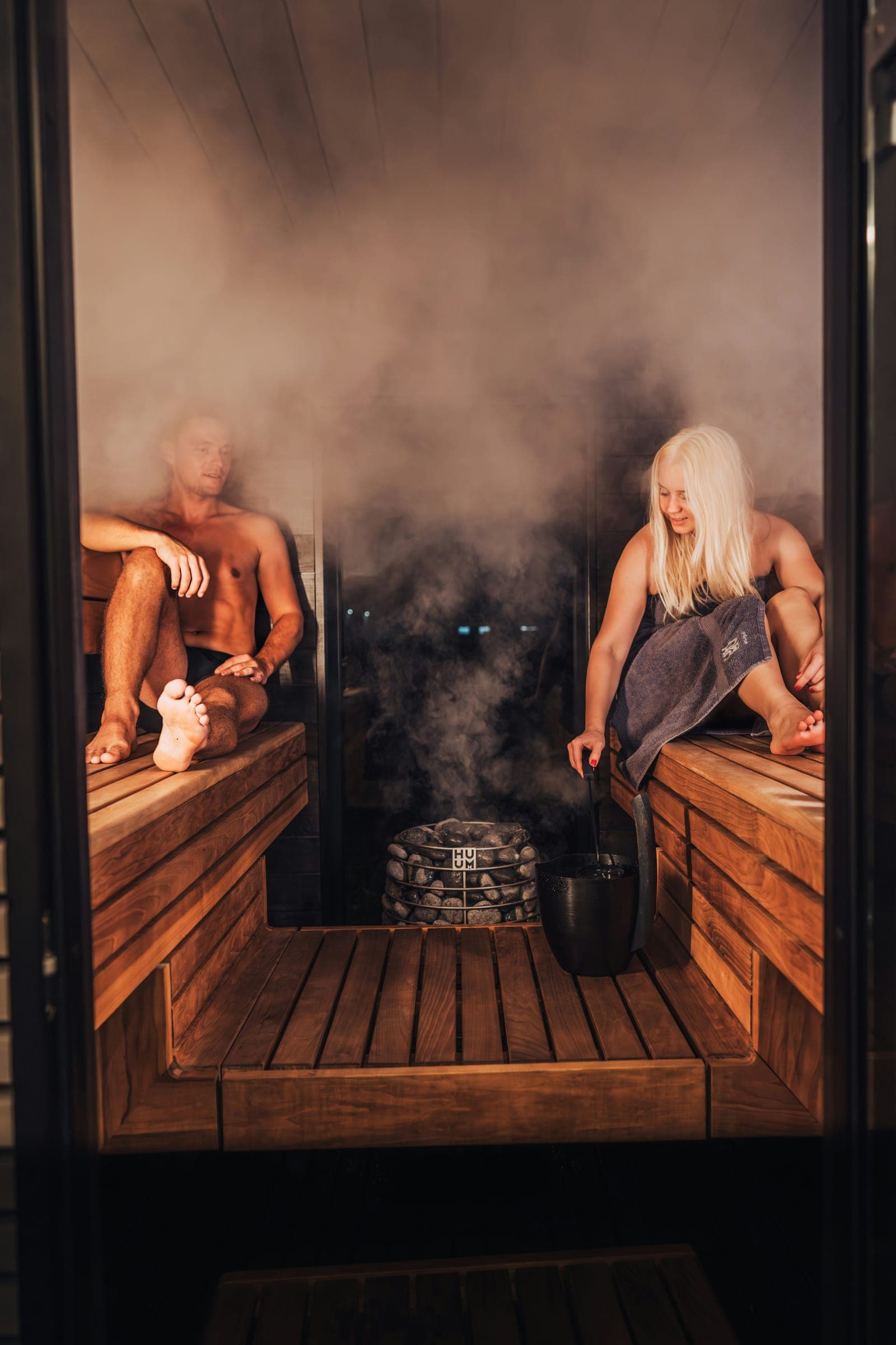 Two people sitting in a steam-filled sauna at night, one reaching for the ladle