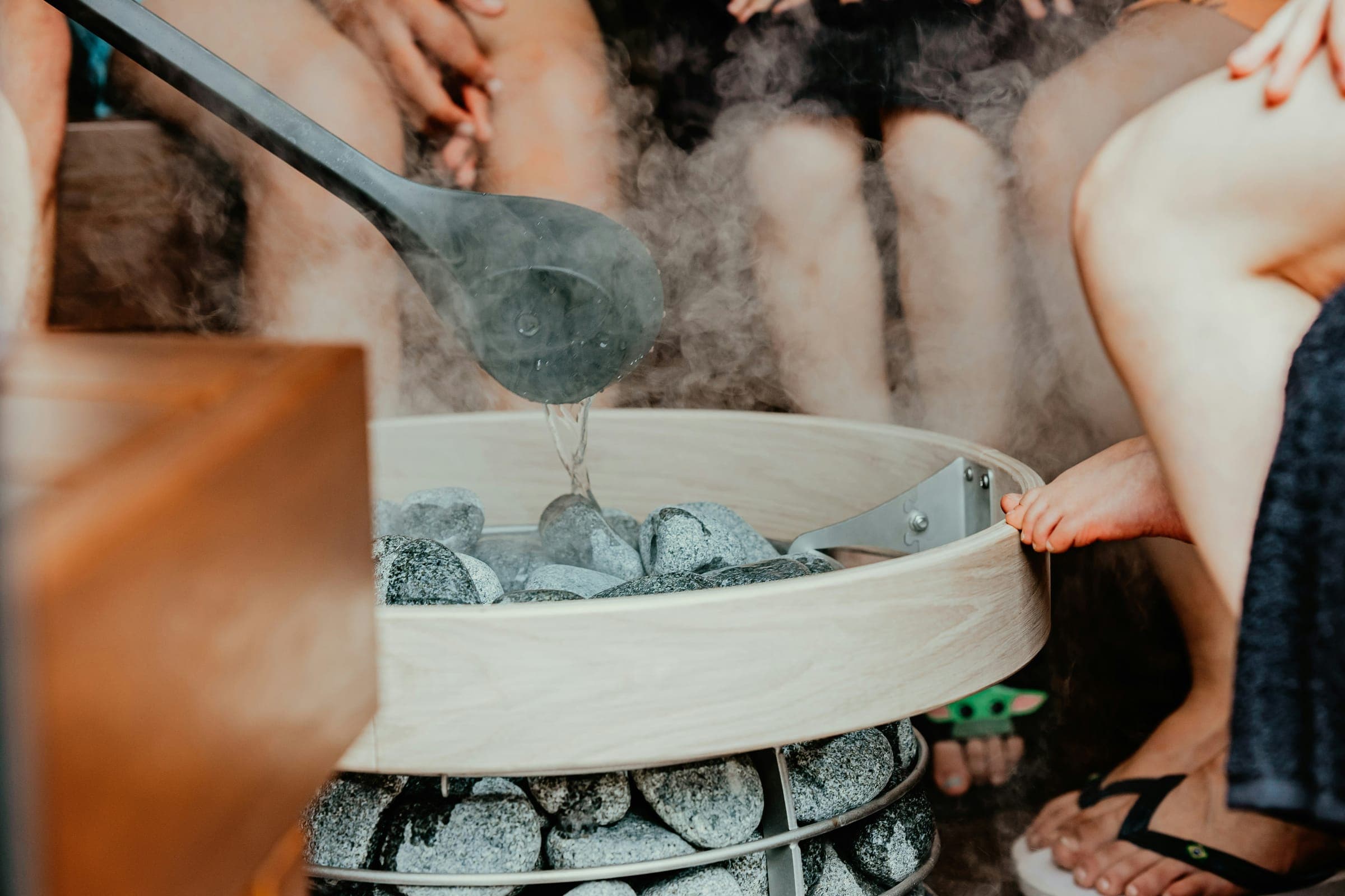 A ladle pouring water over the stones of a sauna heater while several people sit around it