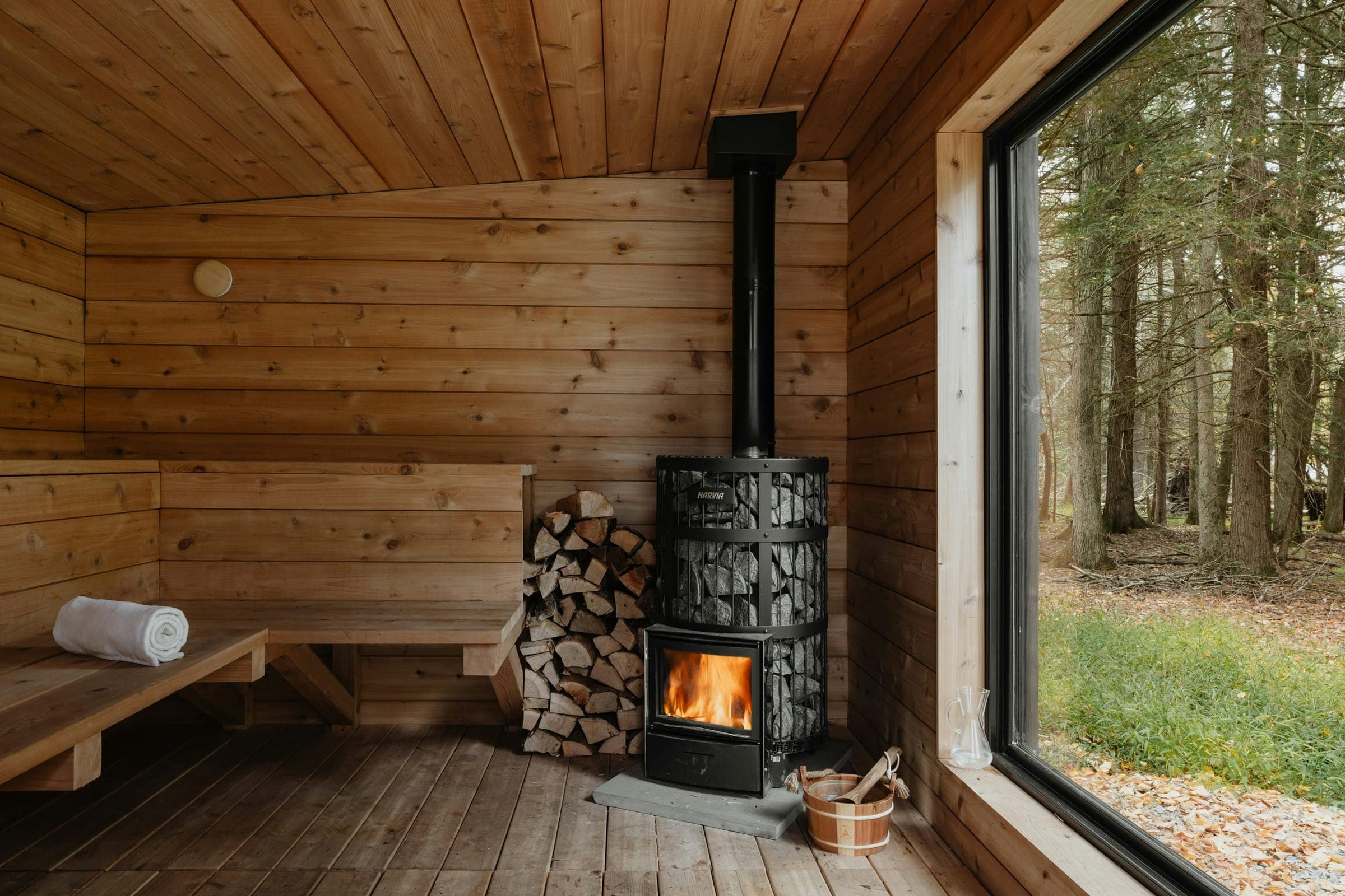 A wood-burning stove and cedar benches inside a sauna, forest visible through a tall window